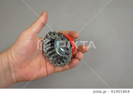 A man's hand holds a bowl wire brush. A tool for woodworking as well as stripping surfaces of corrosion, scale, or rust. Side view. Gray background. Selective focus. A man's hand holds a bowl wire brush. A tool for woodworking as well as stripping surfaces of corrosion, scale, or rust. Side view. Gray background. Selective focus. 91905868