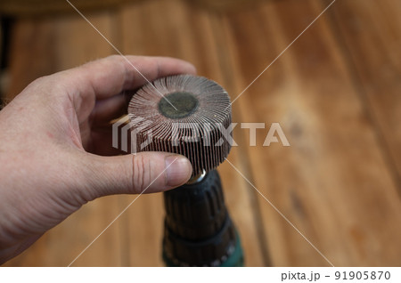 Man places flap wheel in cam of an electric drill chuck. Tool for woodworking, sanding, removing rust from various surfaces. Yellow wooden boards defocused in background. Close-up. Selective focus. 91905870