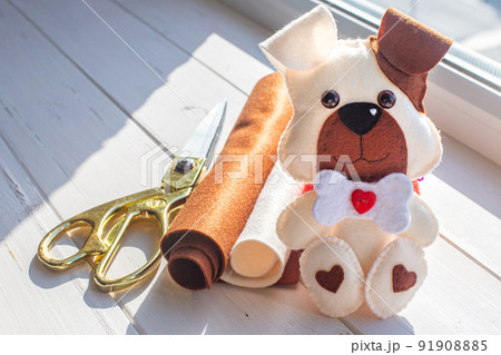 A beautiful puppy made of felt with a brown ear and muzzle, with a bone and a red heart sits on a background of white boards. Tailor's scissors, felt rolls. Creativity, hobby, sewing toys with a child 91908885