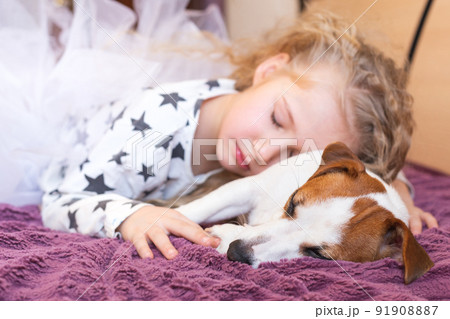 A blonde little girl in pajamas with asterisks and a dog Jack Russell Terrier sleep together in an embrace. The concept of love for pets, friendship, hugs, childhood. Daytime sleep. Day of dogs. 91908887