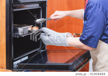 A chef in an apron and protective gloves pulls meat medallions out of the oven with meat tongs. Kitchen, food preparation, cooking. Restaurant, cafe, eatery. Cooking courses, school. 91908889