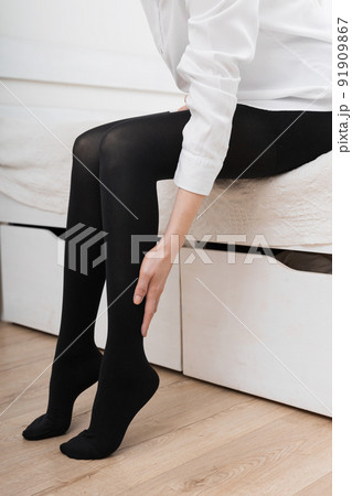 Girl putting on stockings at home. Black compression stockings on a woman in a white room. Black tights. Beautiful female legs. 91909867