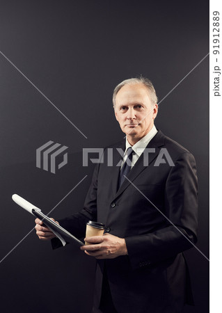 Portrait of content confident owner of business company standing against dark wall and analyzing report in clipboard while drinking coffee 91912889
