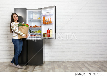 Young woman with paper bag full of products near open refrigerator indoors, space for text 91915556