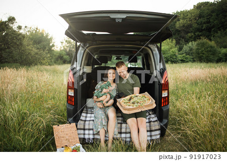 Beautiful and happy family of three sitting in car trunk with tasty pizza on fresh air. 91917023