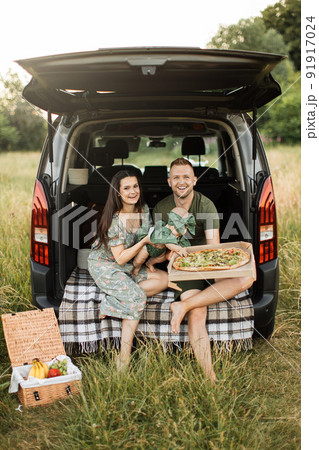 Happy young parents with dog and little daughter unpacking tasty pizza sitting on car trunk outdoors 91917024