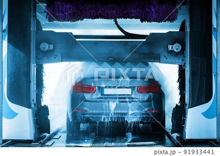 A grey business sedan vehicle running through an automatic car wash with brushes and water hitting car. 91933441
