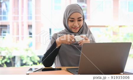 Pleased muslim lady in hijab holding cup of coffee and reading business email or financial information on laptop Pleased muslim lady in hijab holding cup of coffee and reading business email or financial information on laptop 91935006