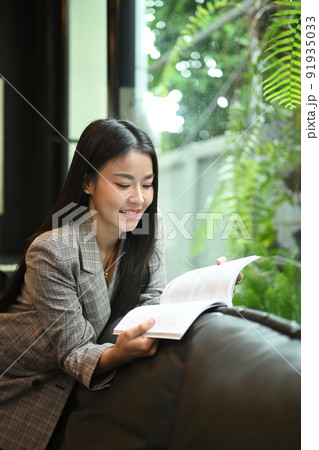 Charming asian woman taking a break from work and reading a book on comfortable couch 91935033