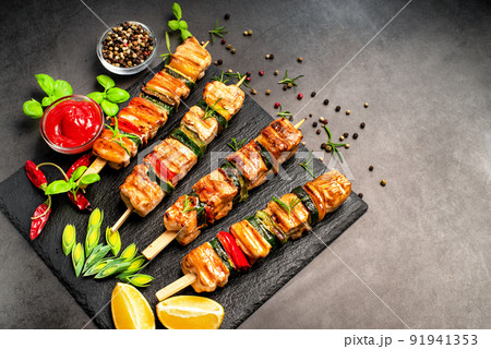 Grilled kebab pieces with vegetables on skewers on a ceramic black board on a dark background with fresh basil leaves.Grilled pieces of chicken meat on skewers. Grilled kebab pieces with vegetables on skewers on a ceramic black board on a dark background with fresh basil leaves.Grilled pieces of chicken meat on skewers. 91941353