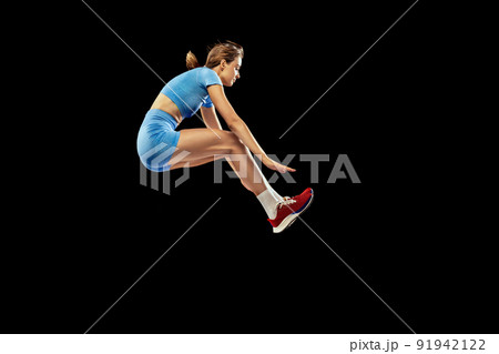 Young sportive girl, long jumper in sports blue uniform performs triple jump isolated on black background. Concept of sport, energy, achievements, motion, speed. 91942122