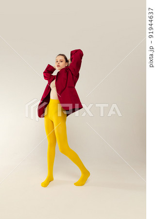 Full-length portrait of young stylish girl in yellow tights and red jacket posing isolated over grey studio background. Bright look Full-length portrait of young stylish girl in yellow tights and red jacket posing isolated over grey studio background. Bright look 91944637