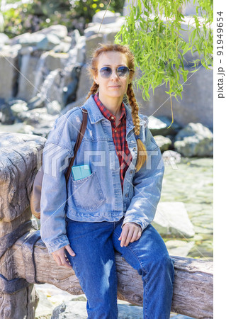 A woman with pigtails in jeans clothes sits on a wooden railing in the shade of a willow. Rest in the spring park. Youth fashion. 91949654