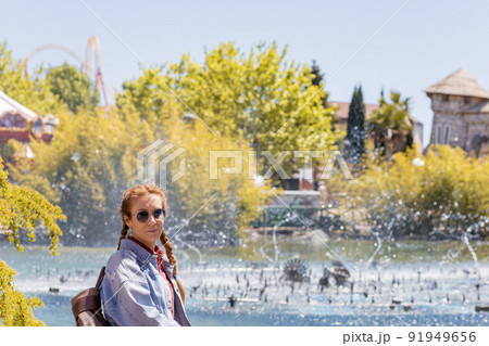 A woman with pigtails stands in front of a fountain gushing from a pond in an amusement park on a sunny day. Spring vacation. 91949656