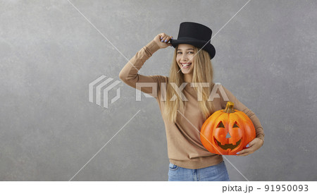 Happy woman in top hat holding Halloween pumpkin standing on grey copy space background 91950093