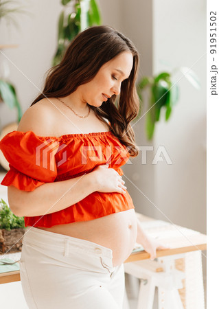 a pregnant woman in a romantic orange blouse and trousers in white interior  91951502