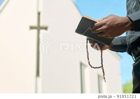 Hands of priest in black apparel holding rosary beads and open Gospel 91951721
