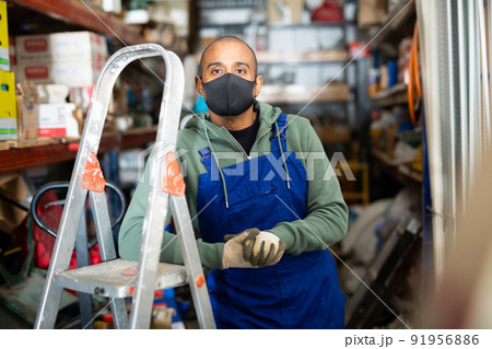 Warehouse worker in protective mask stands next to stepladder and tool shelves in hardware store 91956886