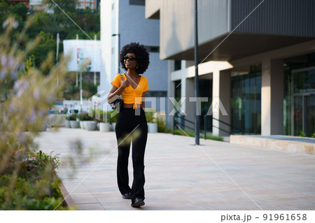 Photo of a stylish young black woman with curly hair wearing orange crop top walking in the street Photo of a stylish young black woman with curly hair wearing orange crop top walking in the street 91961658