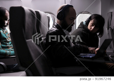 Female passenger looking at man during airplane flight, using laptop online on aerial transportation. Group of tourists travelling by plane on international airline service, aviation jet. 91967246