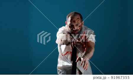 Growling creepy monster prowling at camera on blue background. Mindless looking apocalyptic brain-eating evil monster with deep and bloody wounds acting bizarre and sinister. Studio shot Growling creepy monster prowling at camera on blue background. Mindless looking apocalyptic brain-eating evil monster with deep and bloody wounds acting bizarre and sinister. Studio shot 91967355