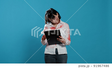 Bizarre looking person with documentation clipboard standing on blue background. Scary dangeorus brain-eating monster with deep and bloody wounds having paperwork documents in hands. Bizarre looking person with documentation clipboard standing on blue background. Scary dangeorus brain-eating monster with deep and bloody wounds having paperwork documents in hands. 91967486