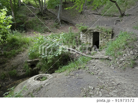 Mysterious stone door in the middle of the forest. Dark portal to the hill among old rocks. Broken trees and moss around. Sunny day. High quality photo. 91973572