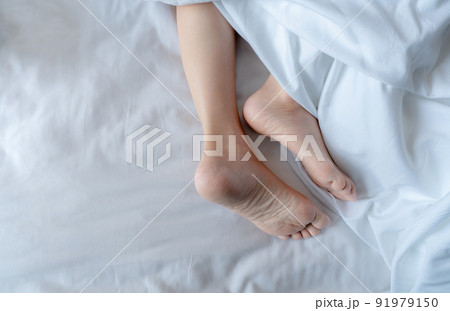 Woman barefoot on bed under white linen blanket in hotel or home bedroom. Healthy sleep and relaxation concept. Lazy Sunday morning. Bare feet of woman chilling sleep on white comfort bed and duvet. 91979150