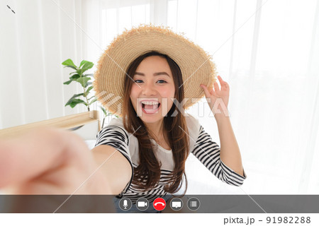 Young Asian woman blogger smiling in summer casual clothes and straw hat talking to facetime video calling with smartphone in bedroom. Using videochat online app with mic, video, phone, share screen, 91982288