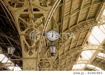 Round Roman clock and glass roof of railway or subway station. Twelve o'clock, seven past twelve pm. 91982864