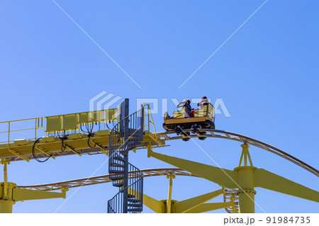 Yellow slide ride with people sitting in a small trailer against the blue sky. Extreme attraction in the amusement park Yellow slide ride with people sitting in a small trailer against the blue sky. Extreme attraction in the amusement park 91984735