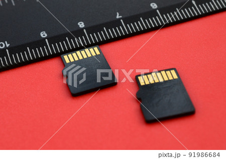 Two small micro SD memory cards lie on a red background next to a black ruler. A small and compact data and information store Two small micro SD memory cards lie on a red background next to a black ruler. A small and compact data and information store 91986684