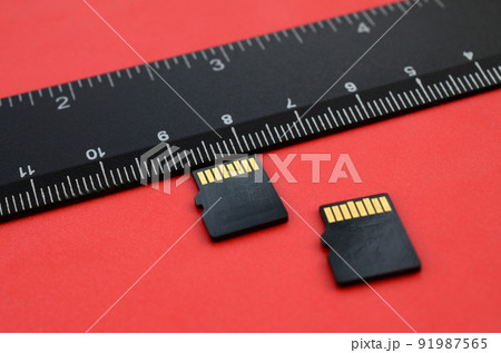 Two small micro SD memory cards lie on a red background next to a black ruler. A small and compact data and information store Two small micro SD memory cards lie on a red background next to a black ruler. A small and compact data and information store 91987565