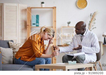 Young bald Afro-American therapist in white coat sitting on chair with clipboard on knees and talking to patient 91989200