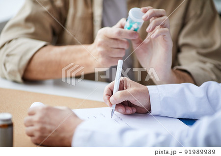 Close-up of unrecognizable male doctor sitting at table and filling prescription while patient reading pill bottle label Close-up of unrecognizable male doctor sitting at table and filling prescription while patient reading pill bottle label 91989989