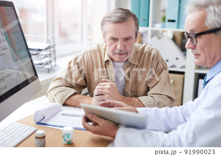 Serious male doctor sitting at desk with computer and showing medical record on tablet to aged patient Serious male doctor sitting at desk with computer and showing medical record on tablet to aged patient 91990023