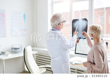 Rear view of gray-haired doctor in lab coat standing against window and watching x-ray image of spinal cord with female patient Rear view of gray-haired doctor in lab coat standing against window and watching x-ray image of spinal cord with female patient 91990310