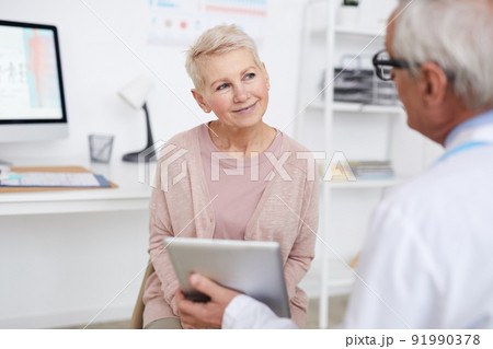 Over shoulder view of elderly doctor using tablet while showing tests results to senior patient at appointment Over shoulder view of elderly doctor using tablet while showing tests results to senior patient at appointment 91990378