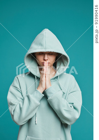 Serious calm young woman in hoodie standing against teal background and holding hands together while praying before fight 91990416