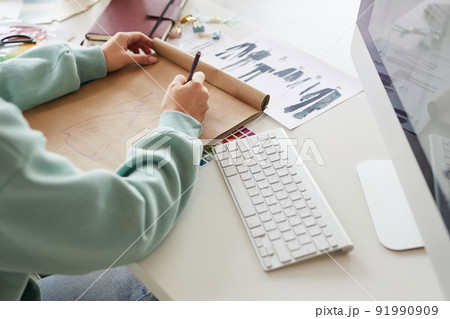 Close-up of unrecognizable clothing designer in hoodie sitting at desk with computer and doing fashion sketch Close-up of unrecognizable clothing designer in hoodie sitting at desk with computer and doing fashion sketch 91990909