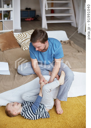 Happy young father in pajamas sitting on bed and tickling sons foot while they having fun together at home Happy young father in pajamas sitting on bed and tickling sons foot while they having fun together at home 91991040