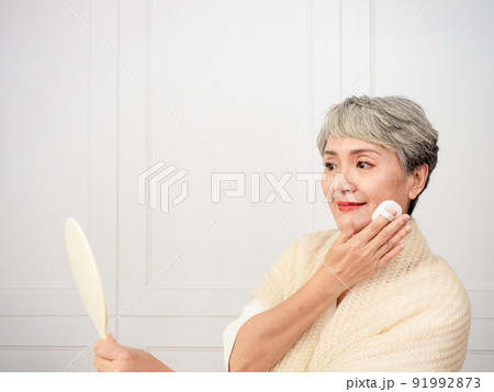 Portrait of mature aged asian woman 50s touching face skin looking in mirror reflection. Smiling mature old lady pampering, healthy moisturized skin care, aging beauty, skincare treatment cosmetics Portrait of mature aged asian woman 50s touching face skin looking in mirror reflection. Smiling mature old lady pampering, healthy moisturized skin care, aging beauty, skincare treatment cosmetics 91992873
