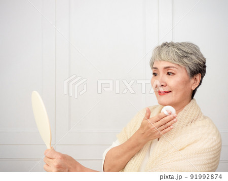 Portrait of mature aged asian woman 50s touching face skin looking in mirror reflection. Smiling mature old lady pampering, healthy moisturized skin care, aging beauty, skincare treatment cosmetics Portrait of mature aged asian woman 50s touching face skin looking in mirror reflection. Smiling mature old lady pampering, healthy moisturized skin care, aging beauty, skincare treatment cosmetics 91992874