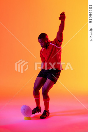 African american male rugby player kicking rugby ball over pink lighting African american male rugby player kicking rugby ball over pink lighting 91994501