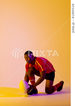 African american male rugby player crouching with rugby ball over pink lighting African american male rugby player crouching with rugby ball over pink lighting 91994526