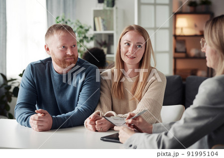 Young couple having an interview with social worker before adoption of child at table during meeting while she making notes in document Young couple having an interview with social worker before adoption of child at table during meeting while she making notes in document 91995104