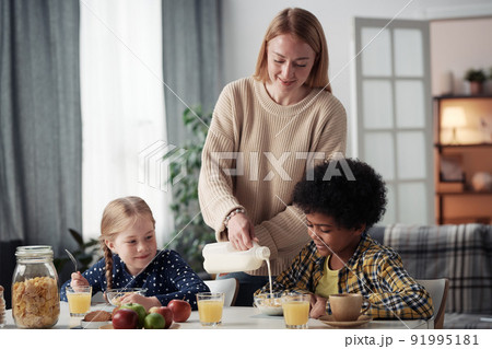 Young caring mom preparing breakfast for her adoptive children, they sitting at table and eating together in the room 91995181