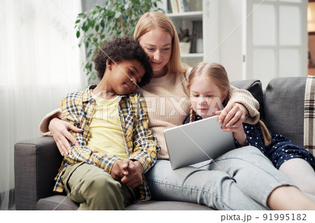 Foster young mother resting on sofa with her adopted children and watching video on digital tablet 91995182