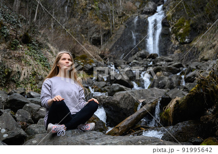 Girl sits on a large stone and meditates near a waterfall next to a mountain river. Girl sits on a large stone and meditates near a waterfall next to a mountain river. 91995642