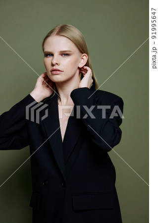 A young charming blonde model tucks her hair behind her ears touches and strokes her curls posing on a green background in the studio A young charming blonde model tucks her hair behind her ears touches and strokes her curls posing on a green background in the studio 91995647